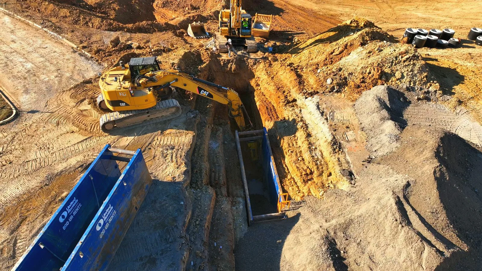 Two excavators digging a utility trench with a blue trench box in it on the graded Downtown Chesterfield site, January 2026