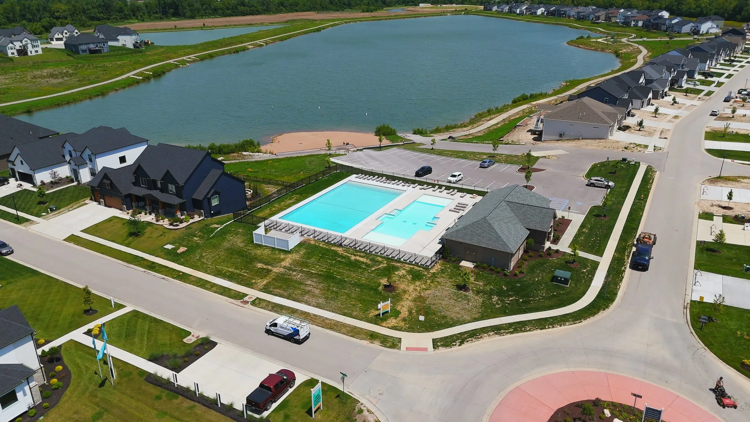 Aerial photograph of Cottleville Trails in May 2026 showing the finished community pool and pool house beside one of the lakes, the sandy beach, and streets of completed homes running away on both sides
