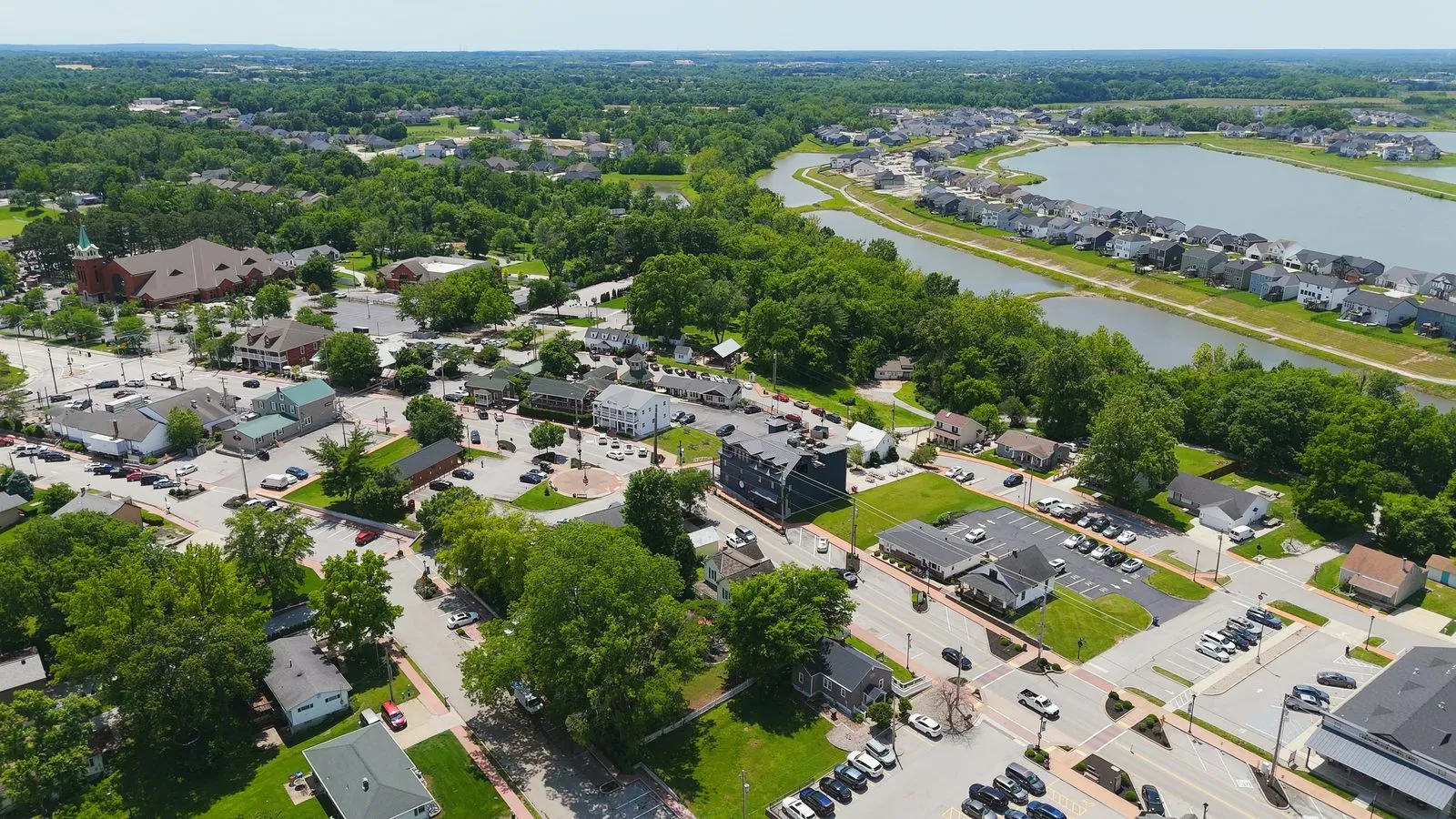 Aerial photograph looking over Old Town Cottleville in May 2026, with the shops and restaurants of the historic district in the foreground and the lakes and homes of Cottleville Trails visible in the background