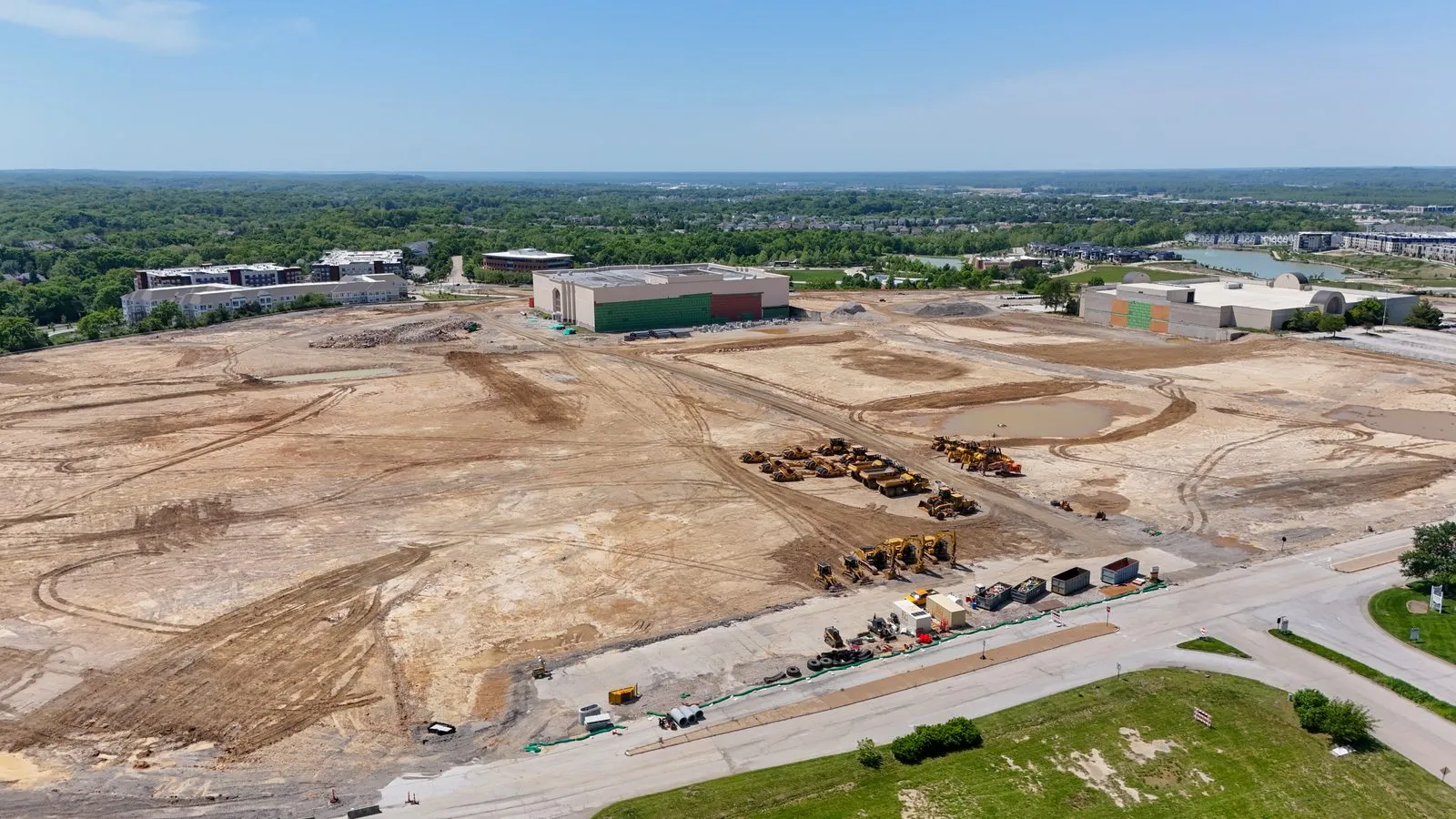 Aerial photograph of the cleared and graded Downtown Chesterfield site in May 2026, with earthmoving equipment lined up along the access road, the retained Dillard’s building at the top of the frame and Wildhorse Village beyond
