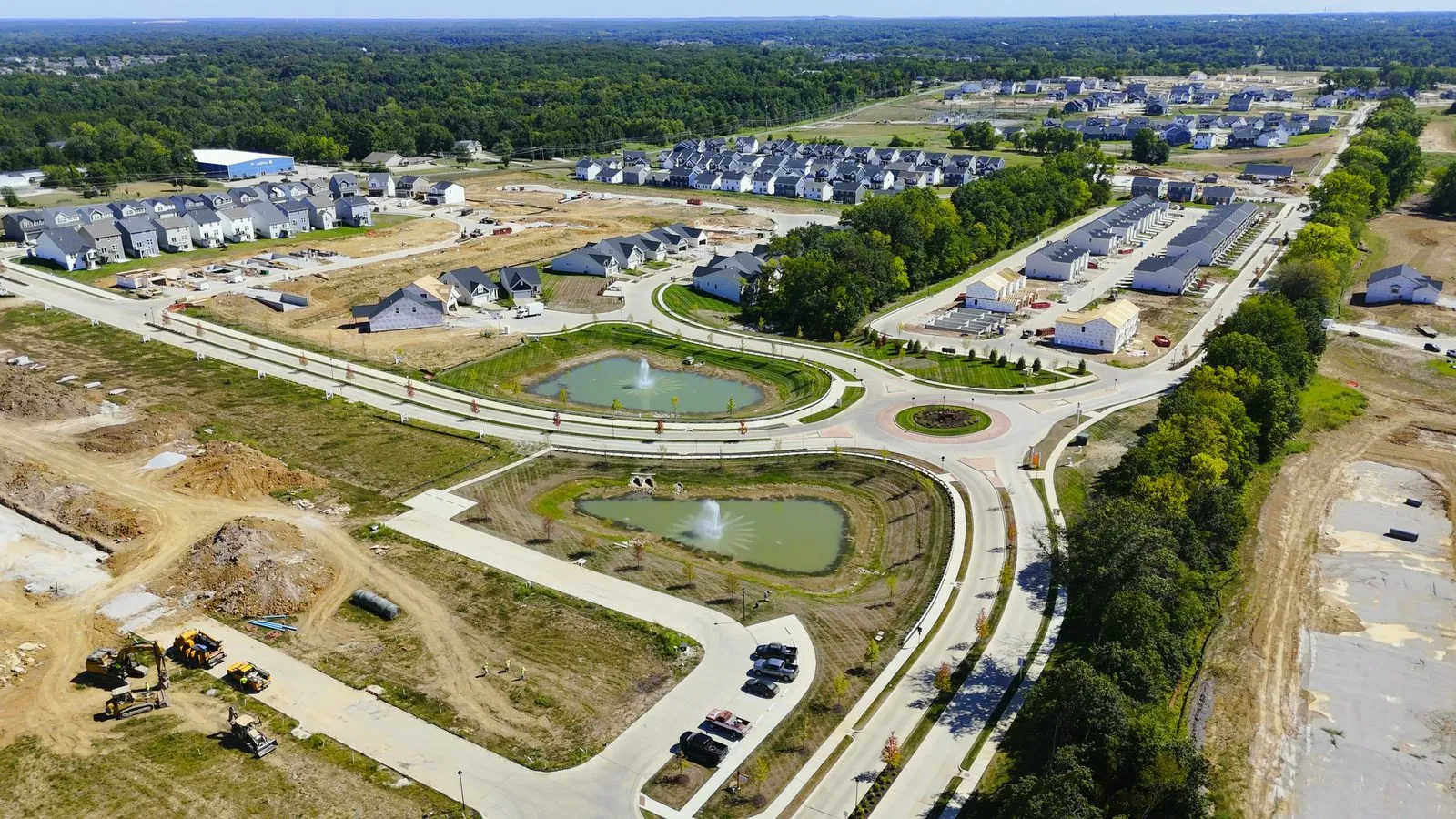 Wide aerial photograph of Harvest in August 2025 showing streets of completed and part-built homes spreading across the site, with graded ground and further villages beyond