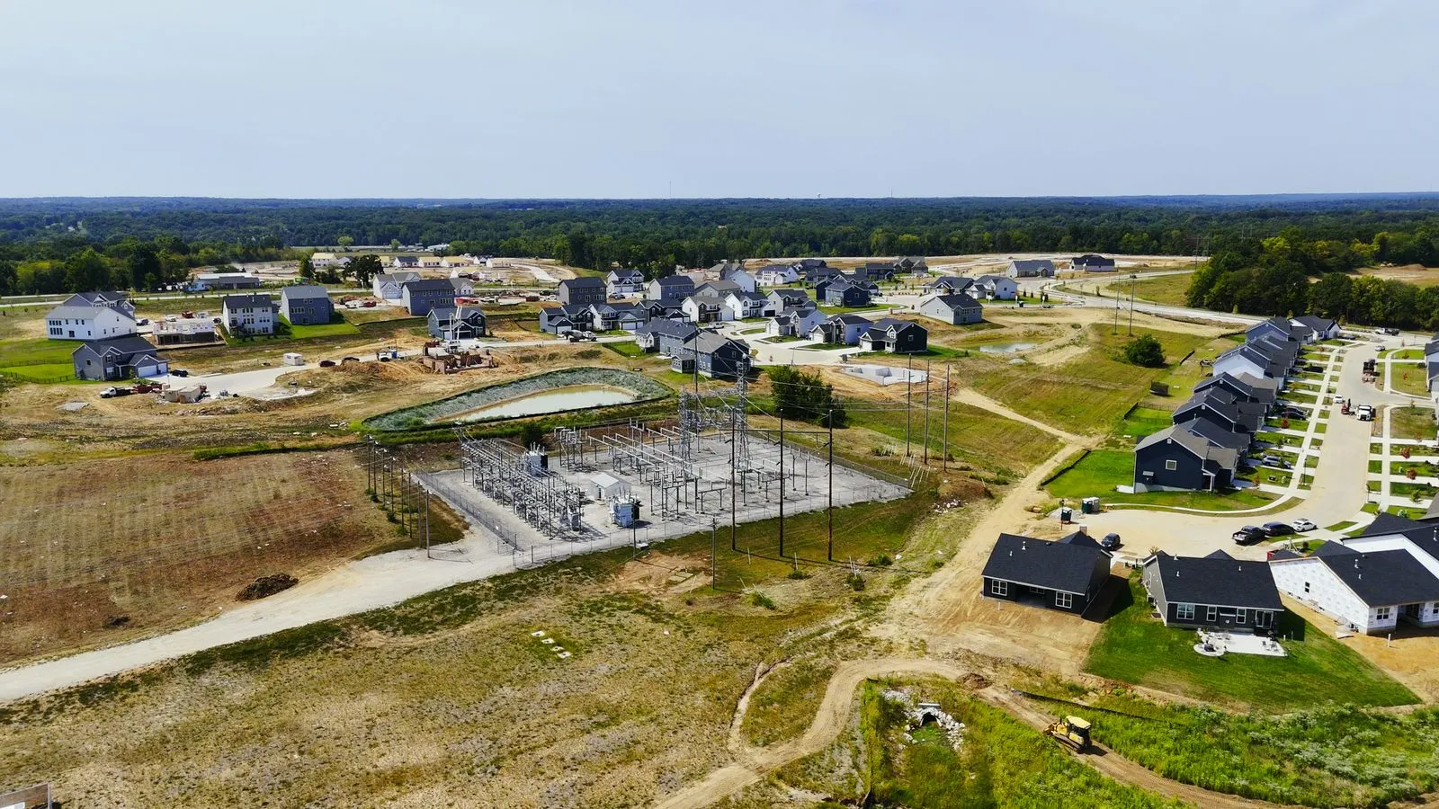 Aerial photograph in August 2025 of the electrical substation beside Harvest, a fenced compound of transformers and steel towers with transmission lines running across the open ground, and completed homes and townhomes immediately to its right