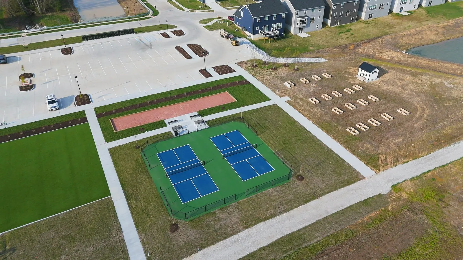 Aerial photograph of the finished town square at Harvest in April 2026 showing two pickleball courts, a bocce court, a civic lawn, a parking lot and rows of raised community garden beds beside one of the lakes