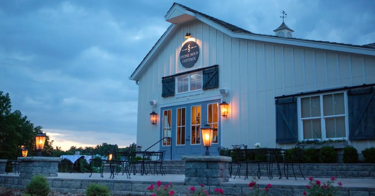 The white farmhouse of Stone Soup Cottage in Cottleville lit up at dusk, with its patio wall and lanterns