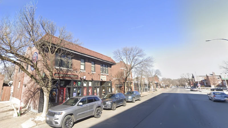 A street-level view along Morgan Ford Road, with parked cars and two-storey brick commercial buildings on both sides