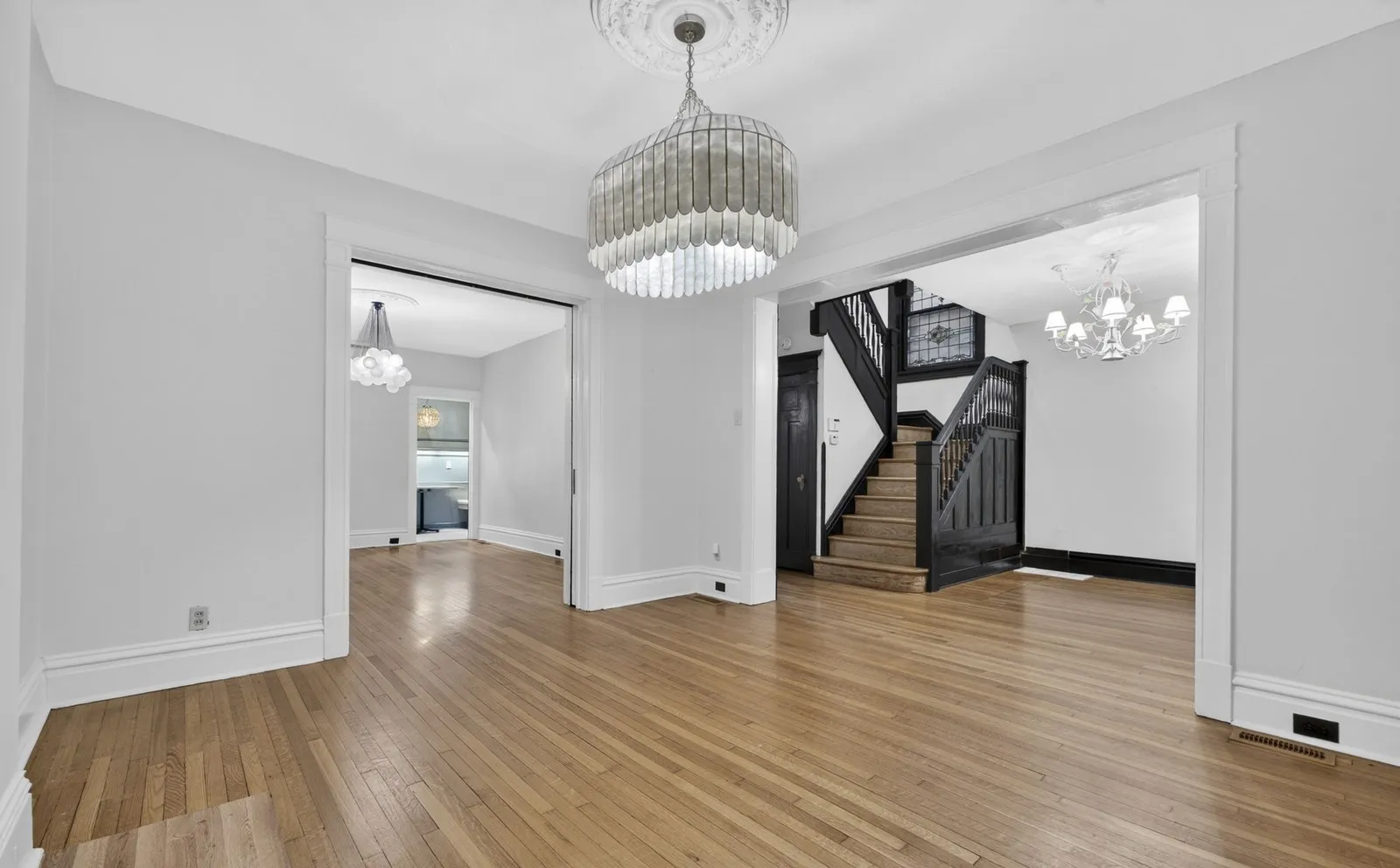 The same dining room empty, with hardwood floors, a chandelier and a black staircase beyond