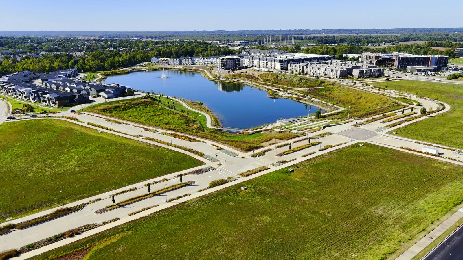 Wide aerial photograph of Wildhorse Village in September 2025 showing the whole 15-acre lake, the modern detached homes on the left, the townhomes and apartment buildings across the water, and large graded parcels in the foreground with streets in but nothing built on them