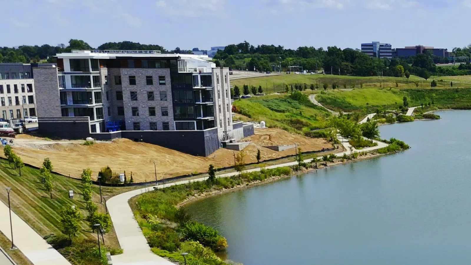 The first Audere condominium building at Wildhorse Village, finished in August 2025, five storeys of stone, glass and balconies standing above the lake with the walking path running past it and the ground around it still bare