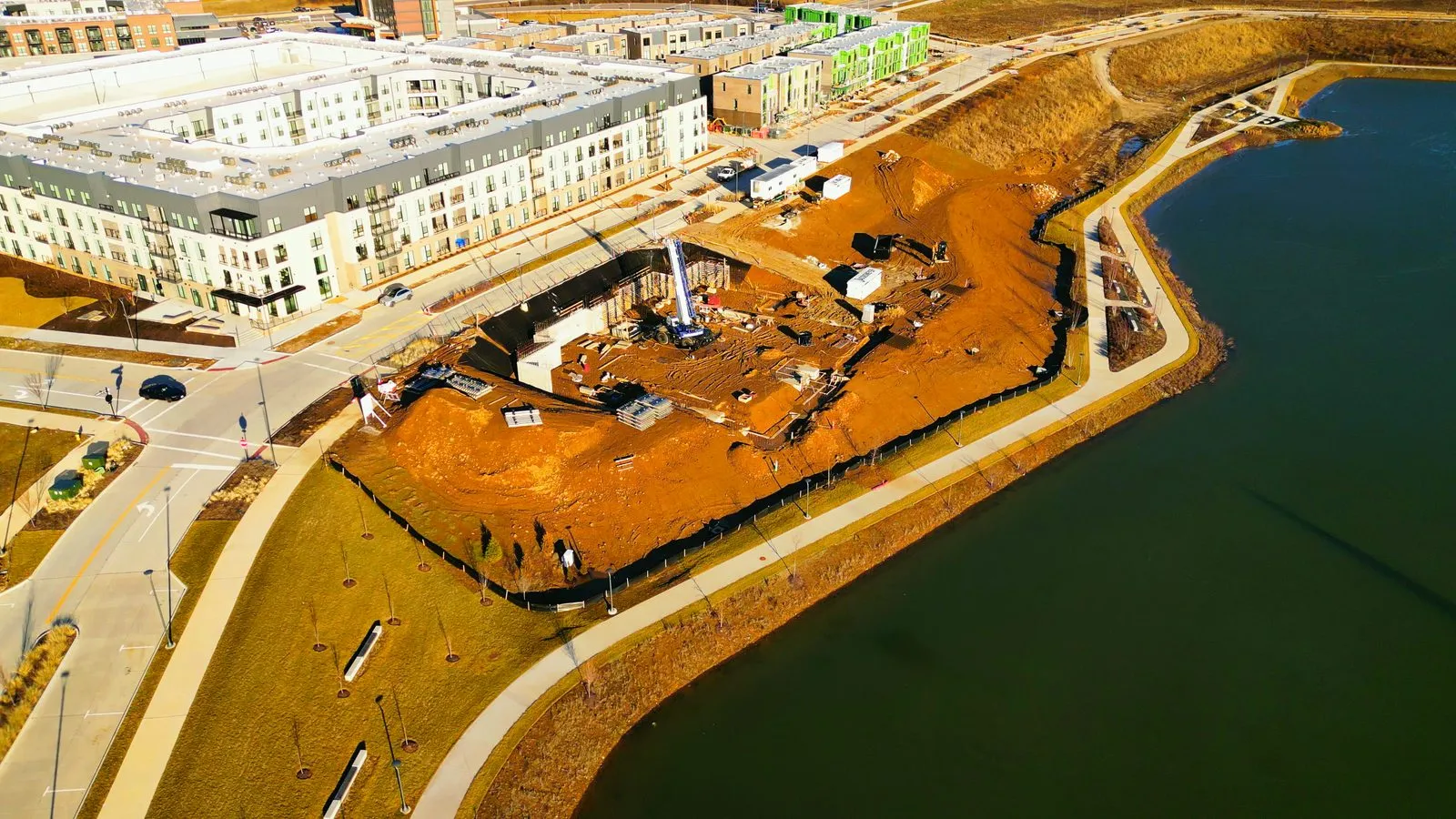Aerial photograph in January 2024 of the excavated foundation for the first Audere condominium building on the lake shore, with the finished Flats apartment building alongside it and rental townhomes in green sheathing behind