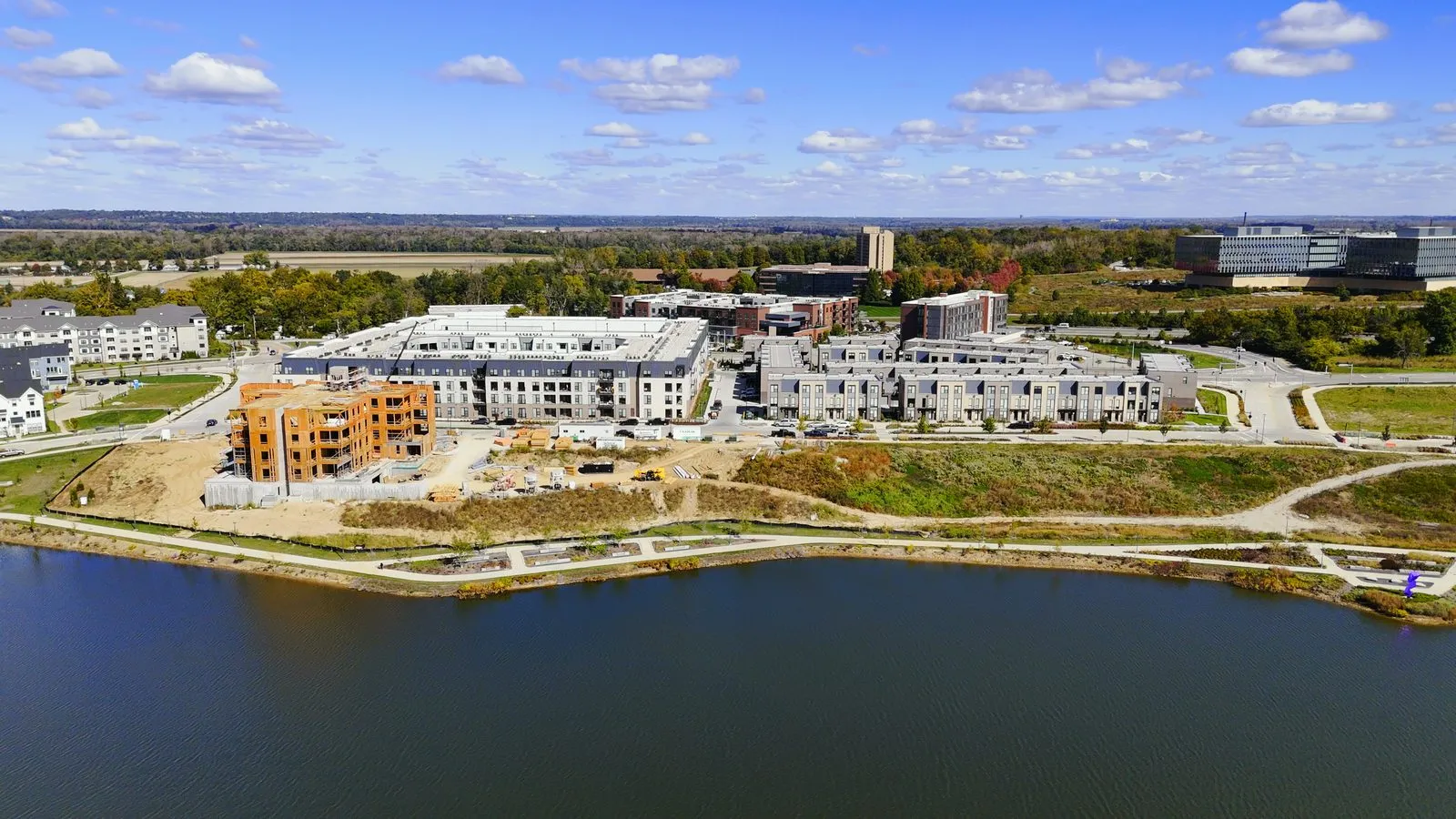 Aerial photograph of Wildhorse Village in October 2024 with the first Audere condominium building in timber framing on the lake shore, the completed Flats apartment building behind it and rows of finished rental townhomes alongside