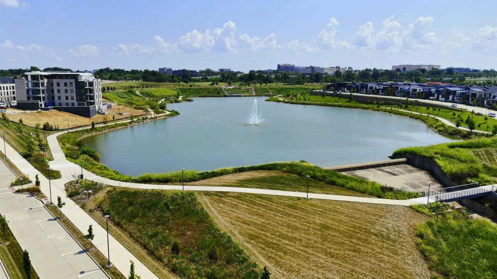 Aerial photograph of the Wildhorse Village lake in August 2025 with the paved walking path running the whole way around it, the finished Audere condominium building on the left and the detached homes on the far shore