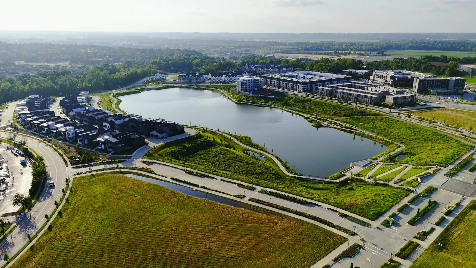 Aerial photograph of Wildhorse Village in June 2025 taken from above the open ground on the south side, showing the lake with the detached homes on one shore and the townhomes and apartment buildings on the other