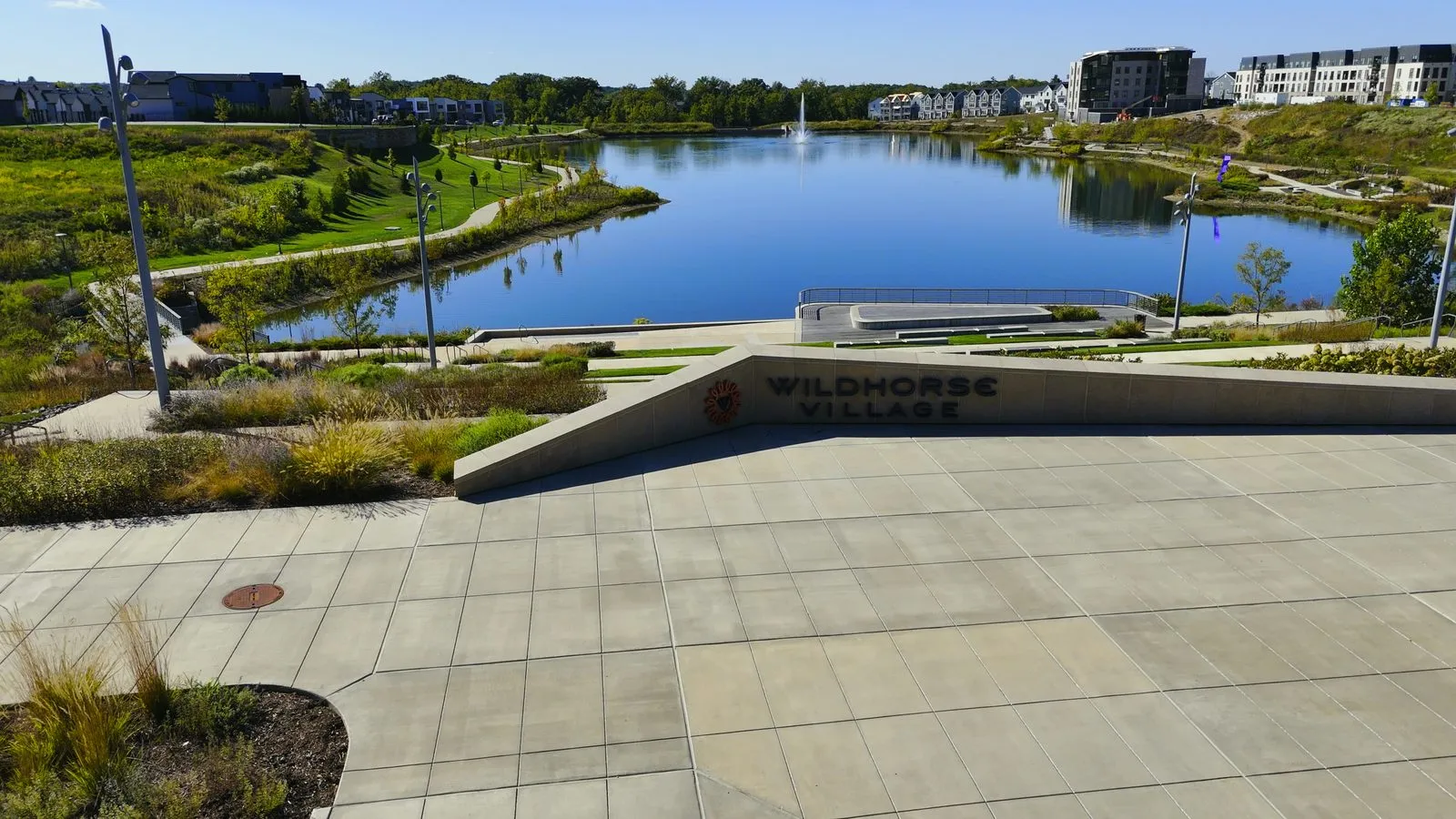The finished terraced plaza at Wildhorse Village in September 2025, with the Wildhorse Village lettering cut into the low stone wall, wide steps dropping to the lakefront boardwalk, and the fountain playing on the water